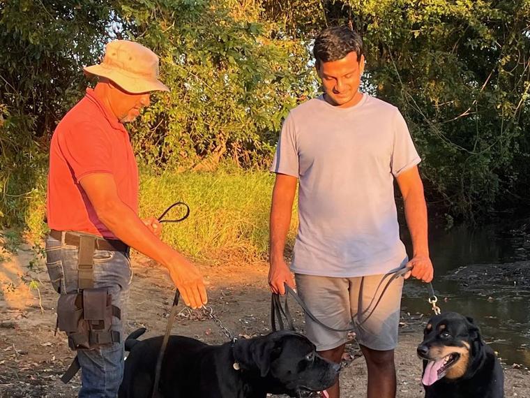 Two handlers standing with two leashed Rottweilers beside a riverbank